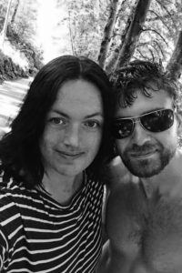 Black and white photo of a Fred & Kjersti standing outdoors on a forest path, smiling at the camera. Kjersti wears a striped shirt, and Fred wears sunglasses with a bare upper body. Sunlight filters through tall trees in the background, creating a peaceful, natural atmosphere.