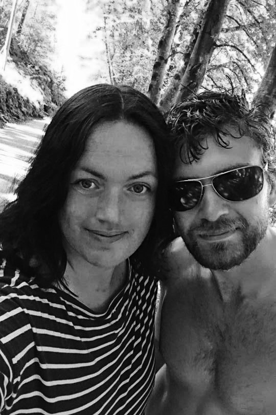 Black and white photo of a Fred & Kjersti standing outdoors on a forest path, smiling at the camera. Kjersti wears a striped shirt, and Fred wears sunglasses with a bare upper body. Sunlight filters through tall trees in the background, creating a peaceful, natural atmosphere.